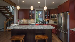 A kitchen with granite countertops, stainless steel appliances, wooden stools, and a view of the outdoors through a window.