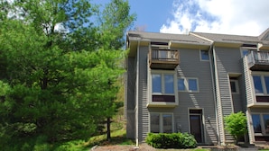 A multi-story residential building with a grey exterior, a front porch, and a small front yard with a tree and shrubbery.