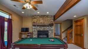 A pool table in a room with a stone fireplace, wooden beams, and a ceiling fan. - Moonrise Retreat 4 Bedroom House (Swanton)