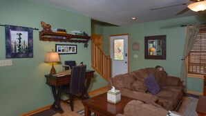 A living room with a brown sofa, a wooden coffee table, a desk with a chair, a lamp, and a ceiling fan.