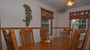 A dining area with a wooden table and chairs, a decorative wall hanging, and windows with red valance curtains. - All About Me 4 Bedroom House (McHenry)