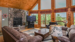 A spacious living room with a stone fireplace, leather sofas, a television, and large windows offering a view of the outdoors.