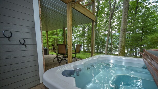 A hot tub with a wooden deck and chairs, surrounded by trees.