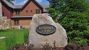 A stone sign with "KENDALL CAMP" engraved on it, in front of a wooden cabin with a stone foundation and a sloped roof. - Summit Lodge 4 Bedroom Townhome (McHenry)