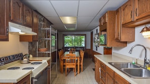 A kitchen with wooden cabinets, a stove, a dining table with chairs, a window with a view of trees, and a sink. - Simpson's Sanctuary 3 Bedroom House (McHenry)