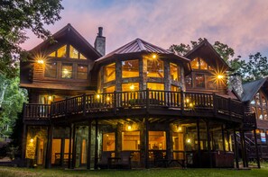 A large, multi-story log cabin with a balcony, large windows, and a stone chimney.