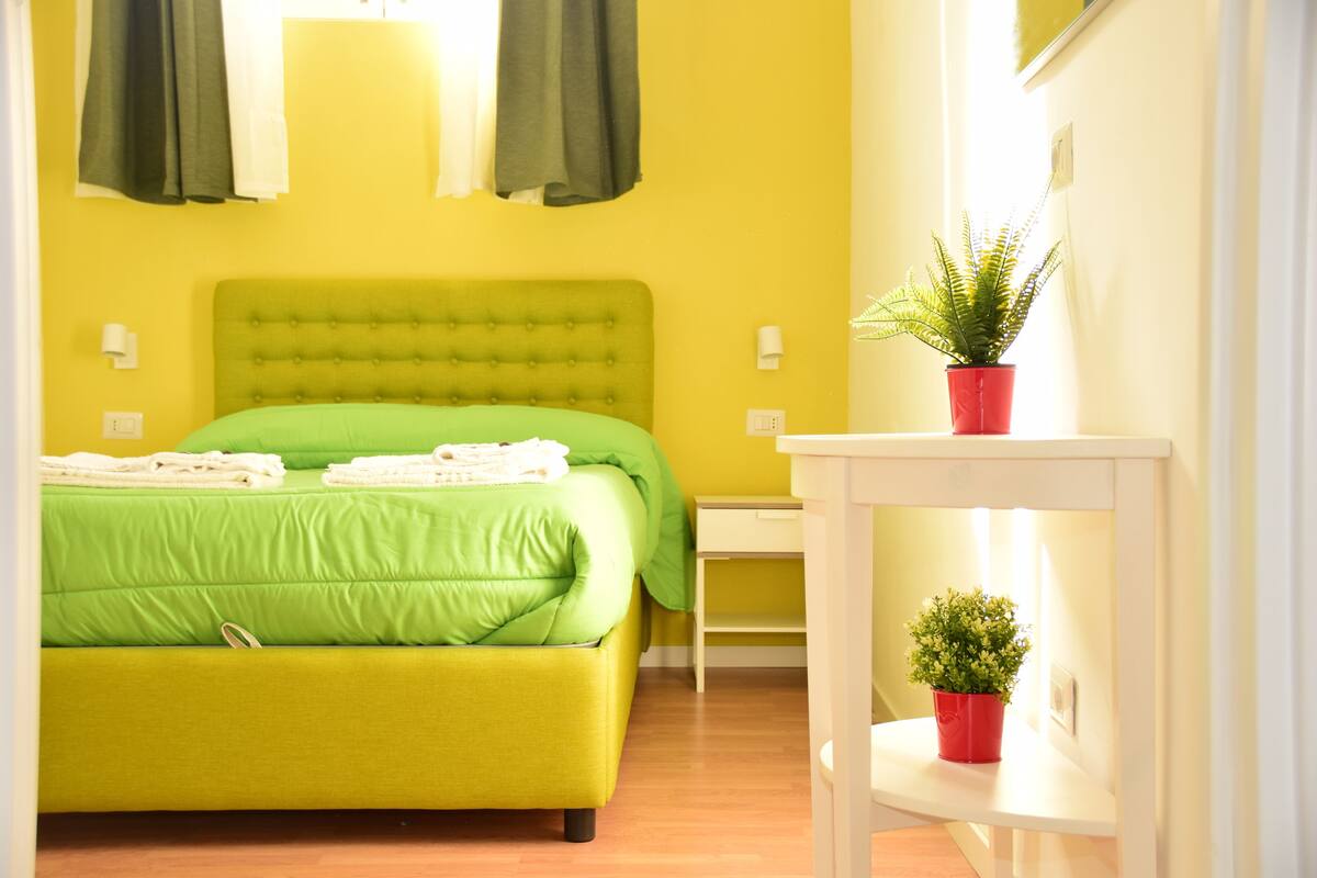 A room with a green bed, a white nightstand, a red potted plant, and a small white shelf.