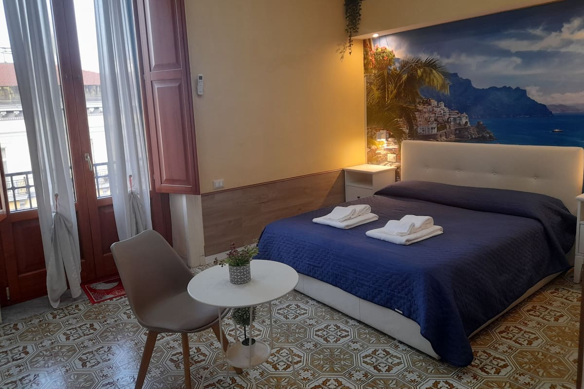 A hotel room with a bed, a chair, a small table with a plant, and a painting of a coastal scene.
