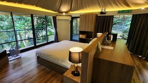 A modern hotel room with a large bed, wooden furniture, a desk with a lamp, and a view of lush greenery outside. - Boulder Valley (George Town)