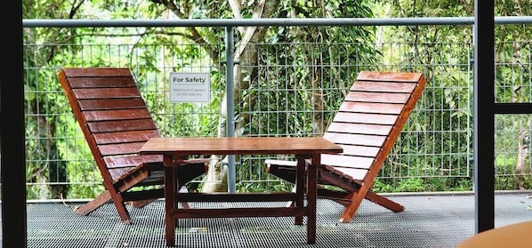 Dos sillones de madera y una mesa en un balcón con baranda de metal, rodeado de follaje.
