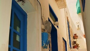 Une rue étroite bordée de bâtiments blancs et bleus, avec un escalier apparent et une petite fenêtre ornée d’une plante.