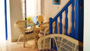 Une pièce avec un escalier bleu, des meubles en osier, un ventilateur de plafond et une fenêtre avec des rideaux.