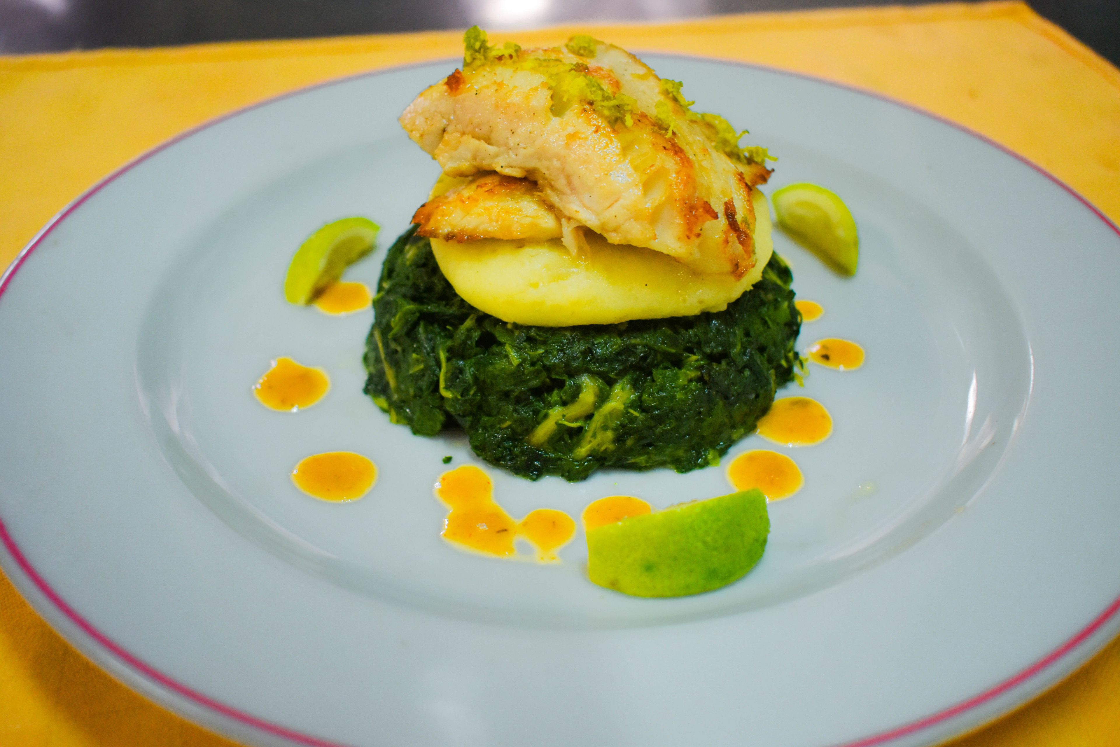 A plate of food with a piece of chicken, a slice of potato, and greens, possibly spinach, on a blue plate with a yellow rim.