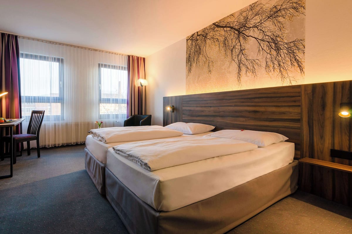 A hotel room with two beds, a desk with a chair, and a large window with curtains.
