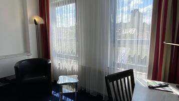 A hotel room with a large window, a desk with papers and a pen, a chair, a small table, and a lamp.