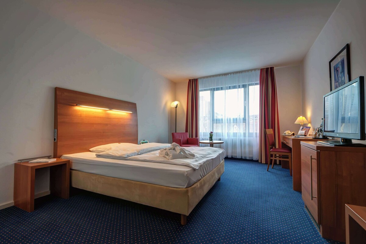 A hotel room with a bed, bedside table, lamp, desk, chair, television, and a window with curtains.