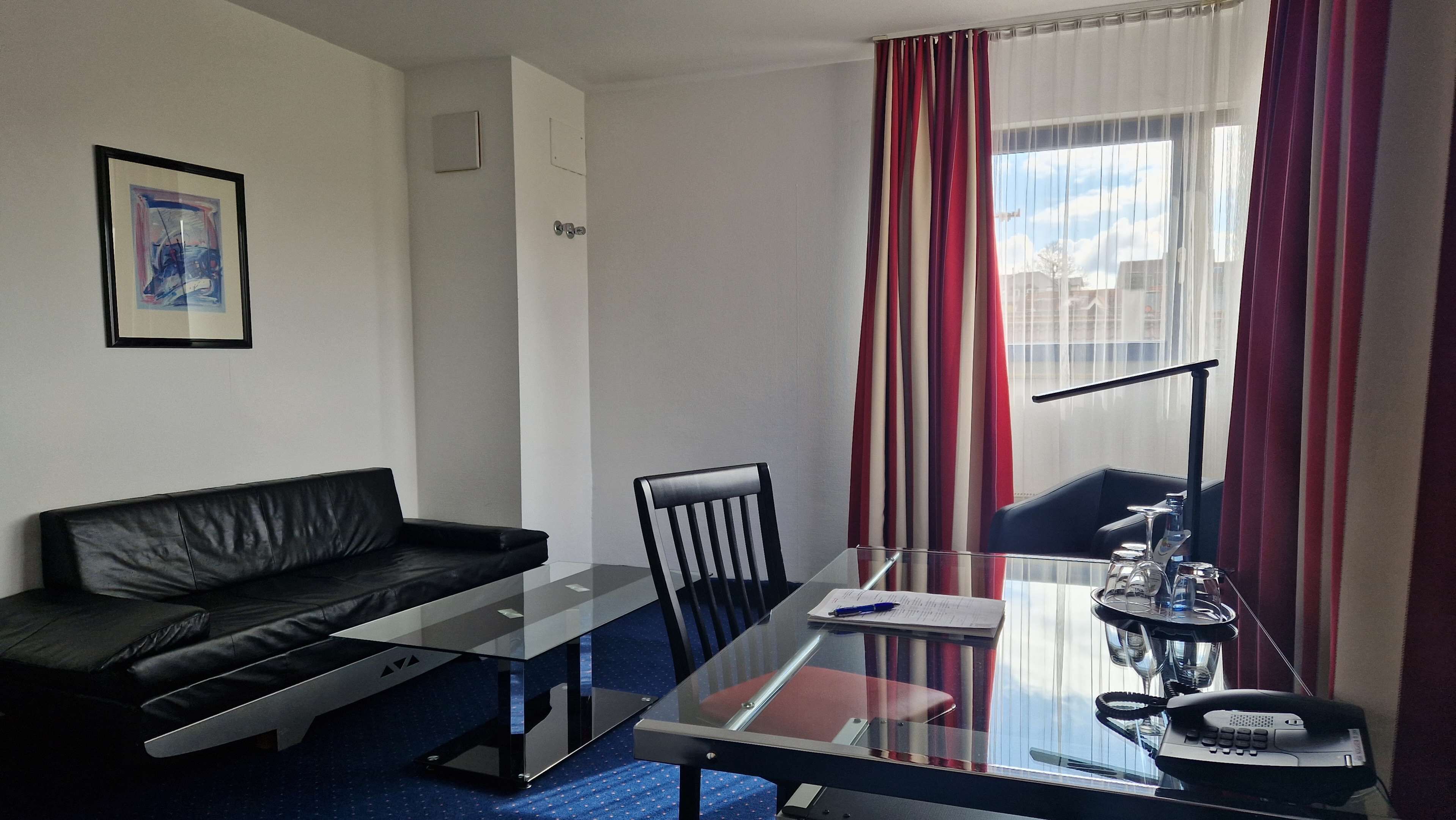 Ein modernes Hotelzimmer mit Glastisch, schwarzem Ledersofa, Stuhl, Schreibtisch mit Telefon und Fenster mit roten Vorhängen.