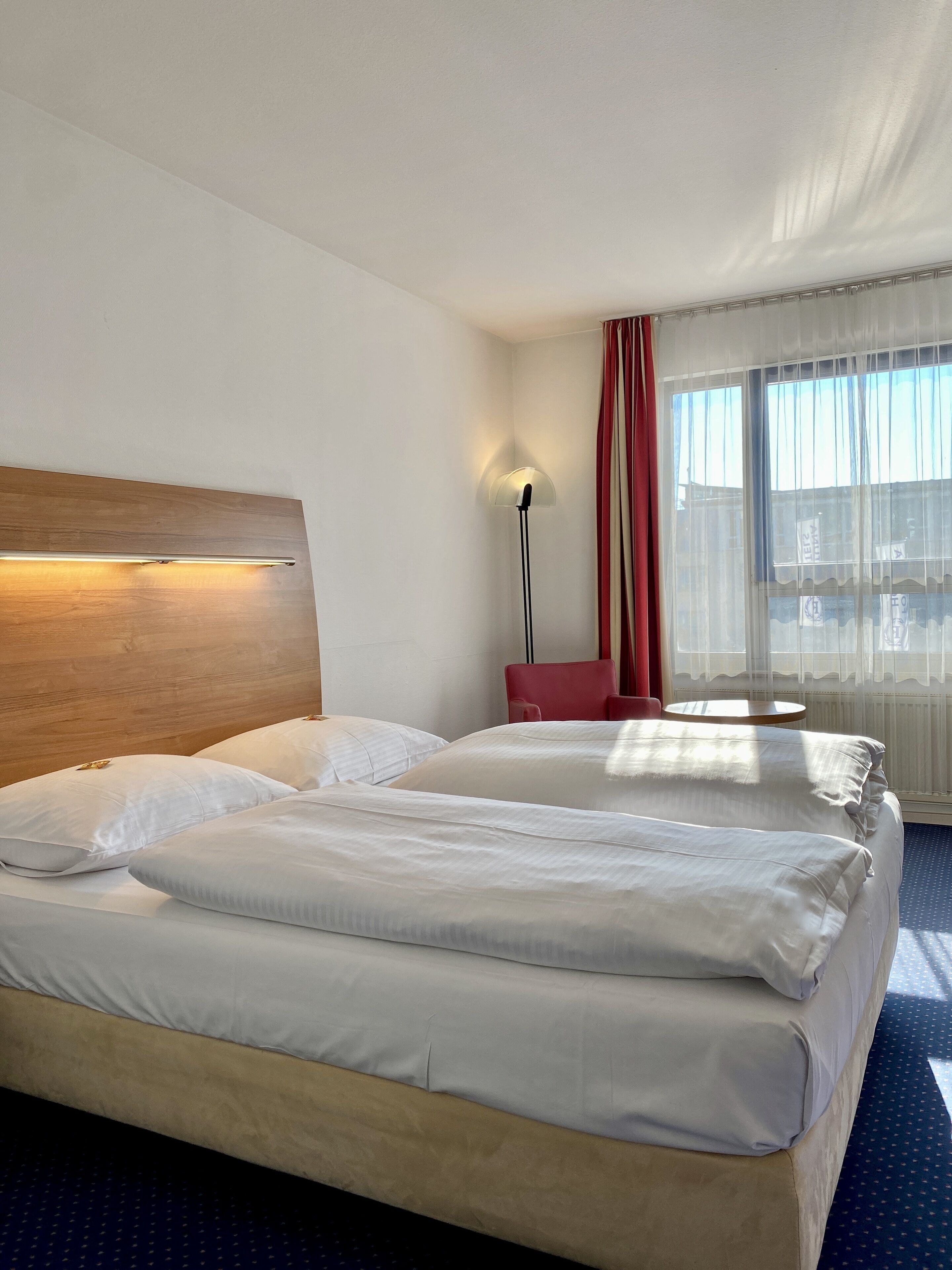Habitación de hotel con dos camas, cabecero de madera, lámpara de pie, una silla roja y una ventana con cortinas translúcidas.