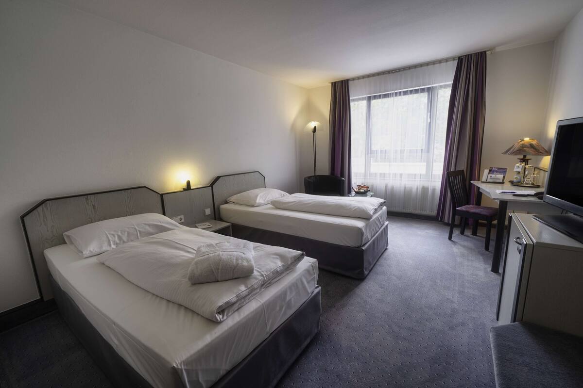 A hotel room with two beds, a desk, a chair, a television, and a window with curtains.