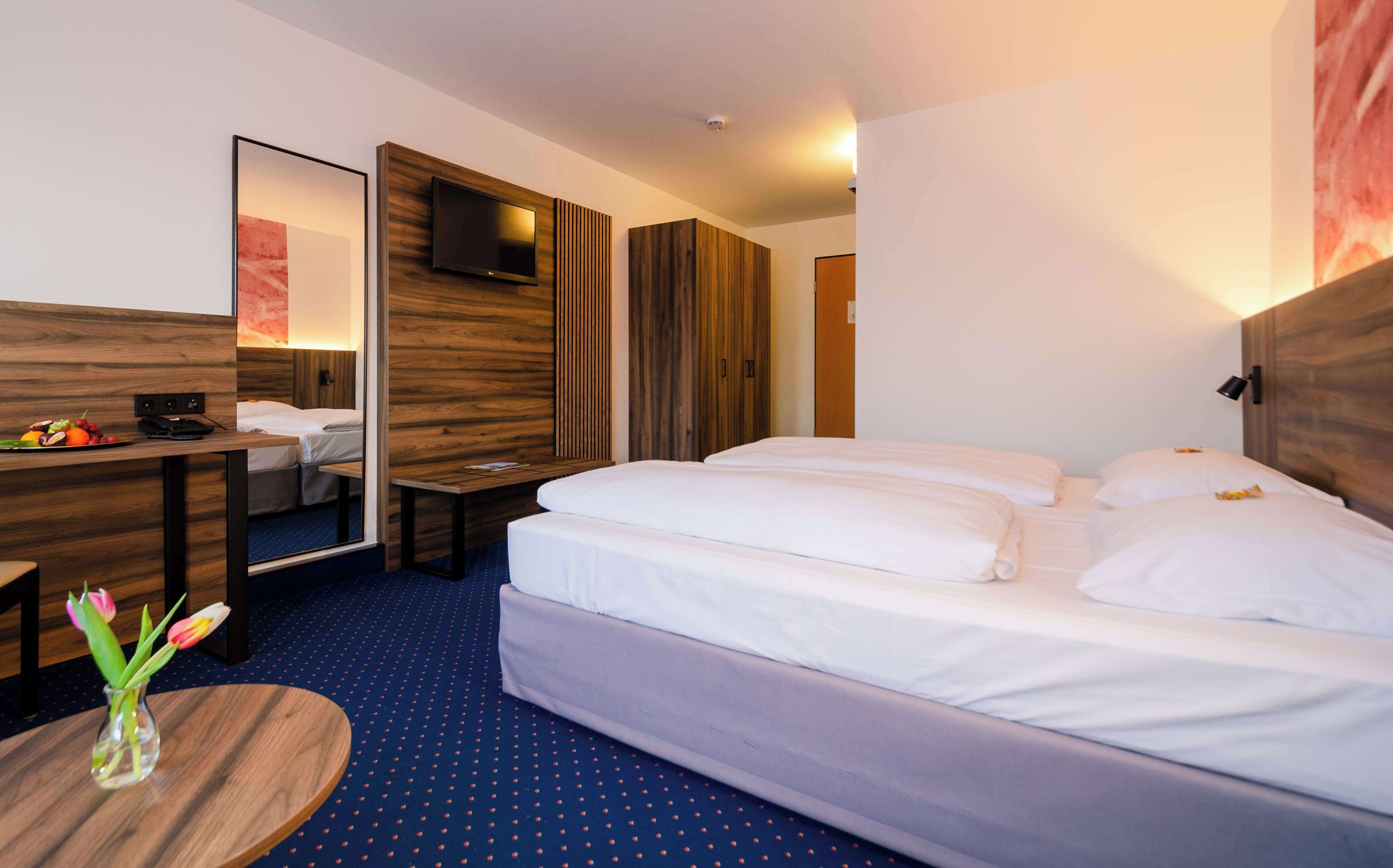 Habitación de hotel con una cama grande, un escritorio con espejo, un televisor montado en la pared y una mesita con un jarrón de flores.