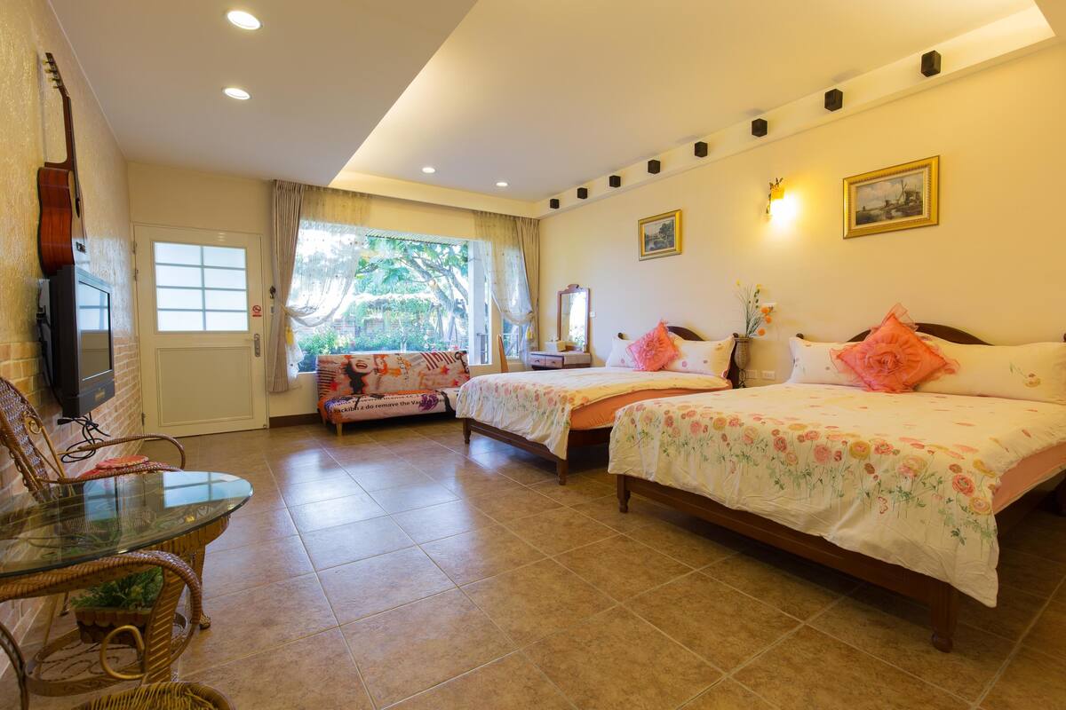 A hotel room with two beds, a glass table, a chair, a TV, and a window with curtains.