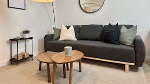 Una sala de estar moderna con un sofá gris, un espejo redondo, una lámpara de pie y una pequeña mesa de madera.