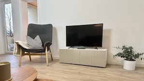 Una sala de estar moderna con un sillón gris, un televisor de pantalla plana sobre un mueble blanco, una mesa de centro de madera y una planta en maceta.
