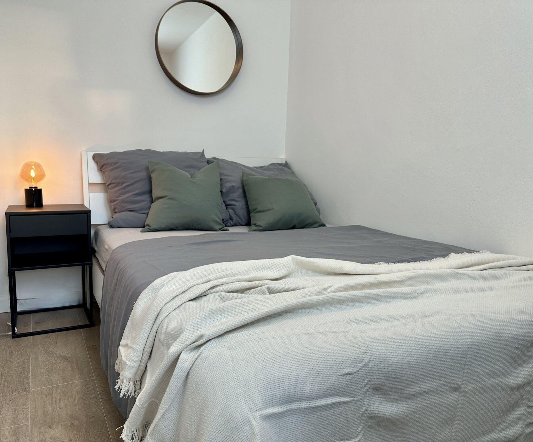 A bedroom with a bed, pillows, a bedside table with a lamp, and a round mirror on the wall.