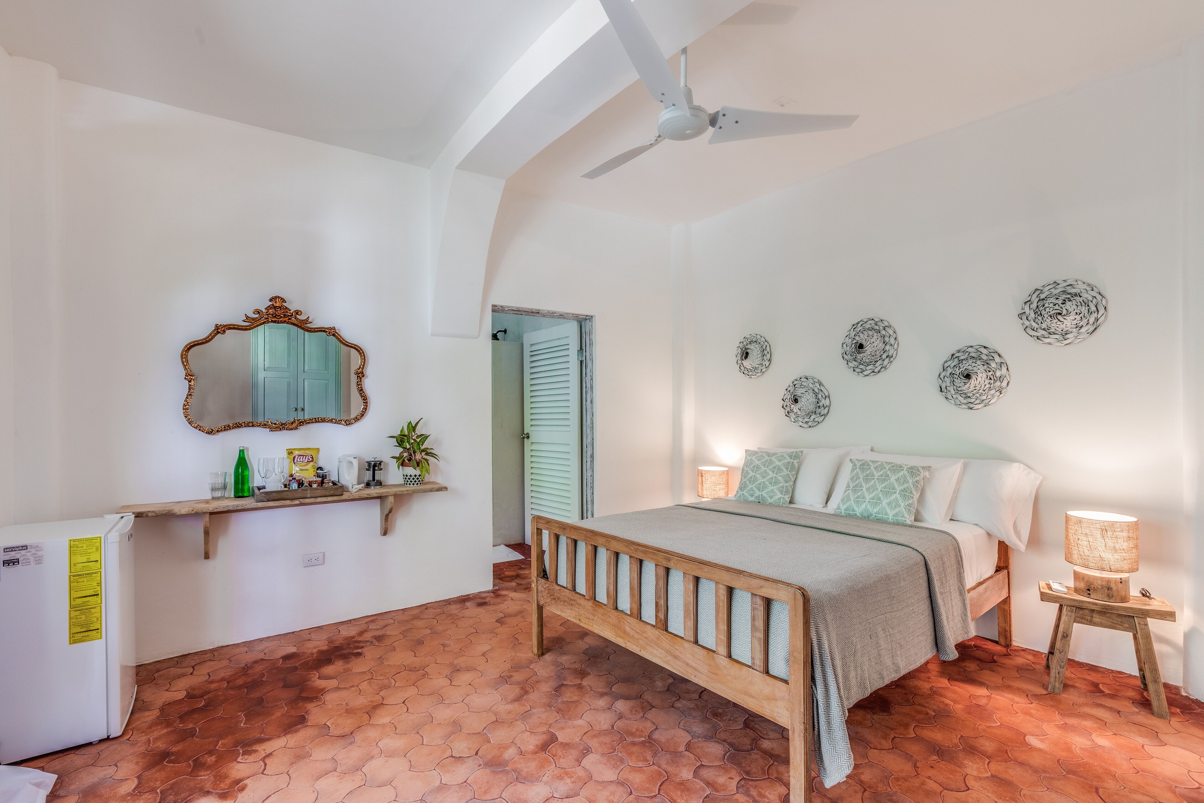 A bedroom with a wooden bed, a ceiling fan, a bedside table with a lamp, a mirror, and decorative plates on the wall.