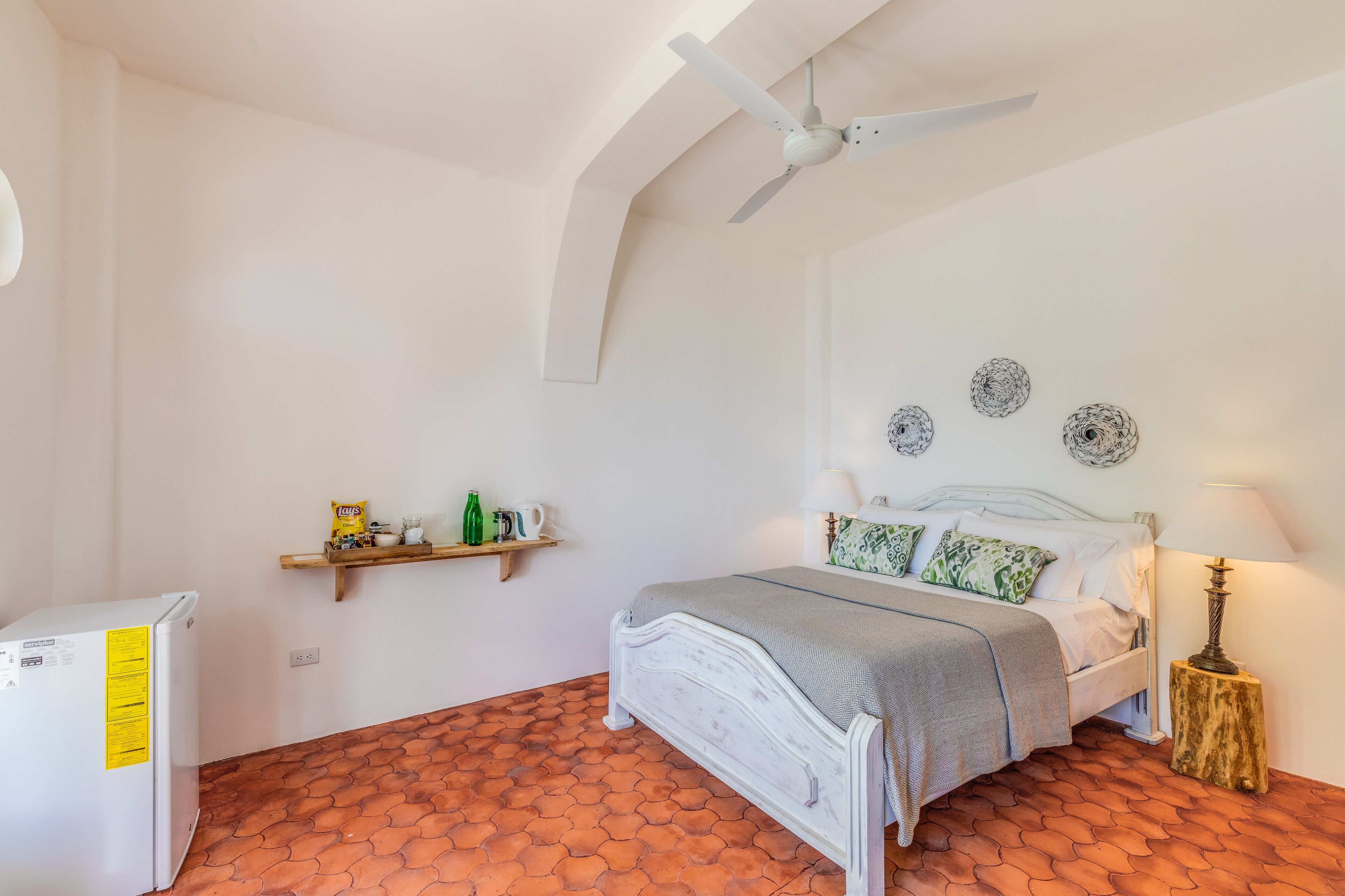 Un dormitorio con una cama blanca, un ventilador de techo, un estante con aperitivos y un refrigerador blanco.