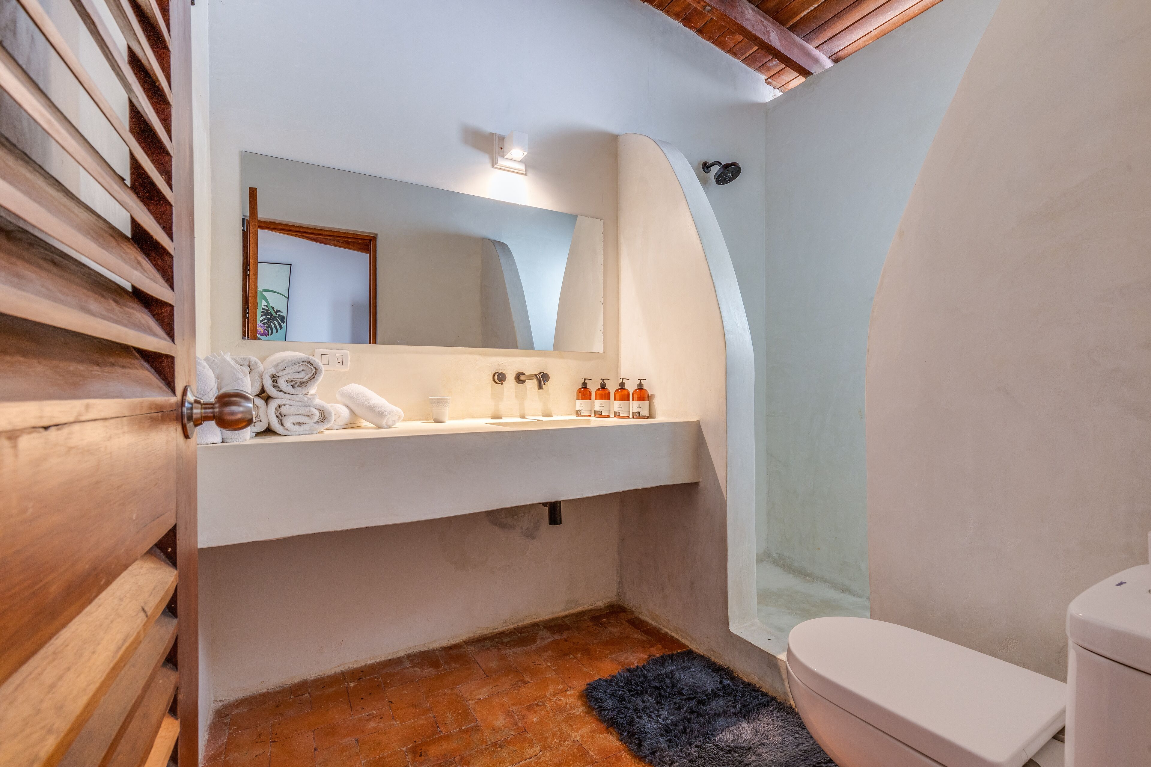 A bathroom with a white sink, mirror, and toilet. There is a towel rack with towels, a black rug, and a wooden bench.