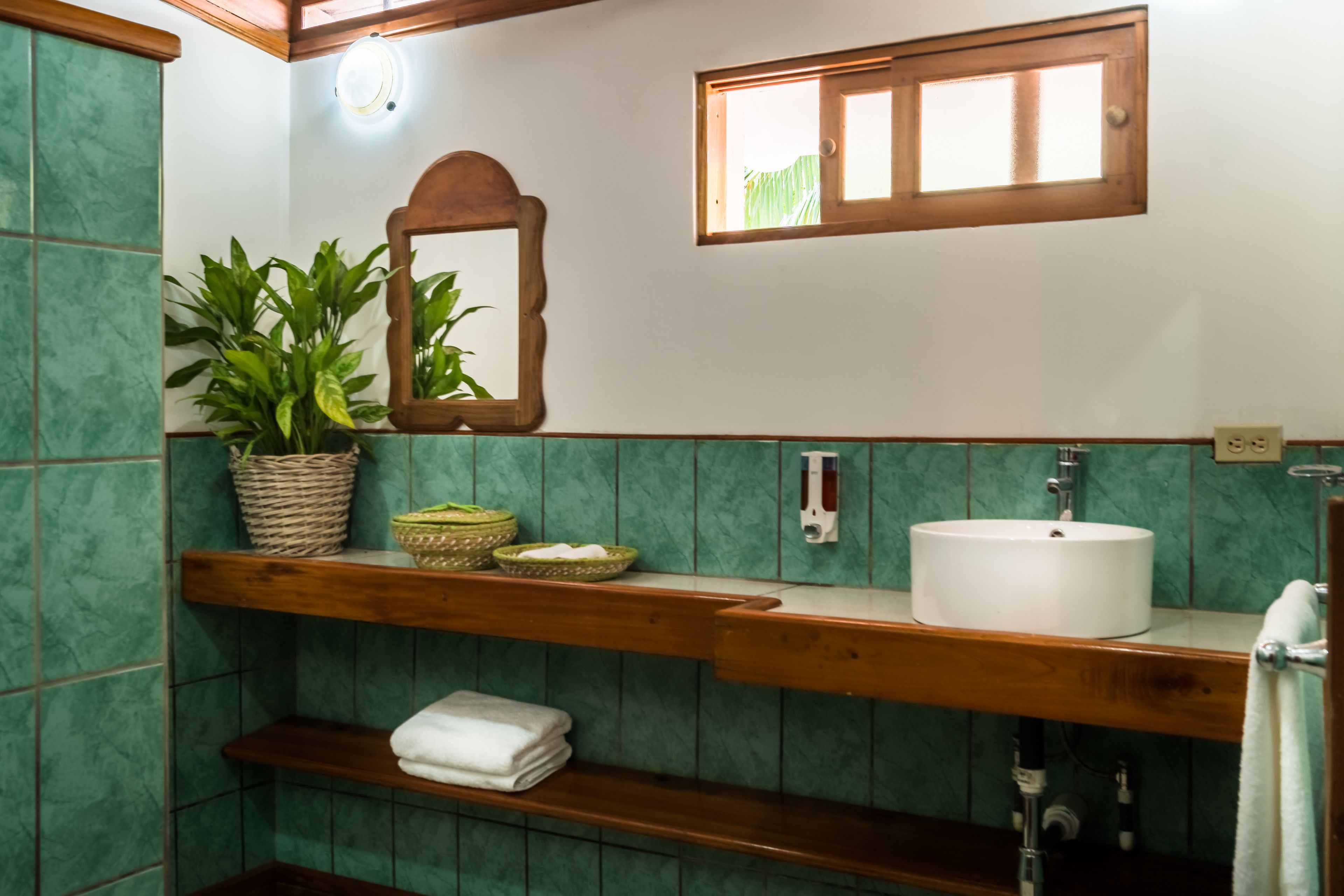 Un baño con pared de azulejos verdes, un estante de madera con toallas, un lavabo blanco, un espejo y una ventana con persianas.