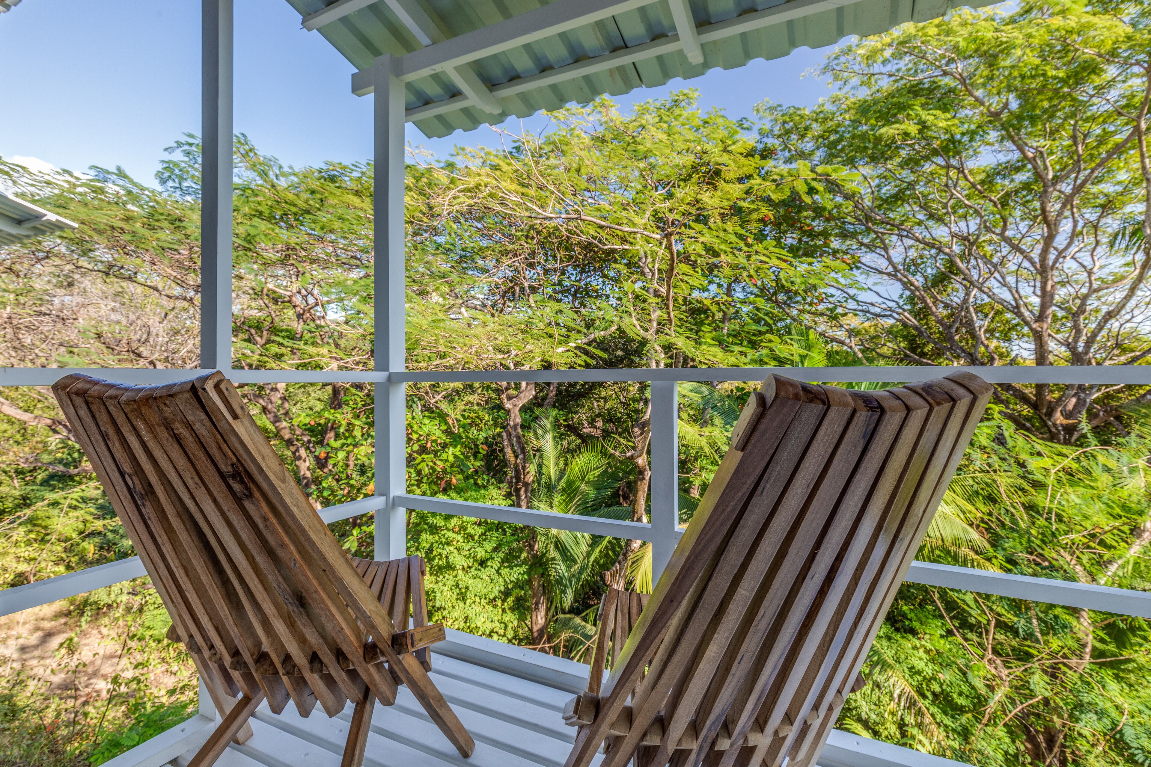 Two wooden lounge chairs on a white balcony with a view of dense green foliage and trees.