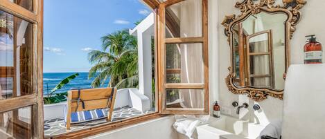 A bathroom with a view of the ocean, a wooden bench, a large ornate mirror, and a white sink.