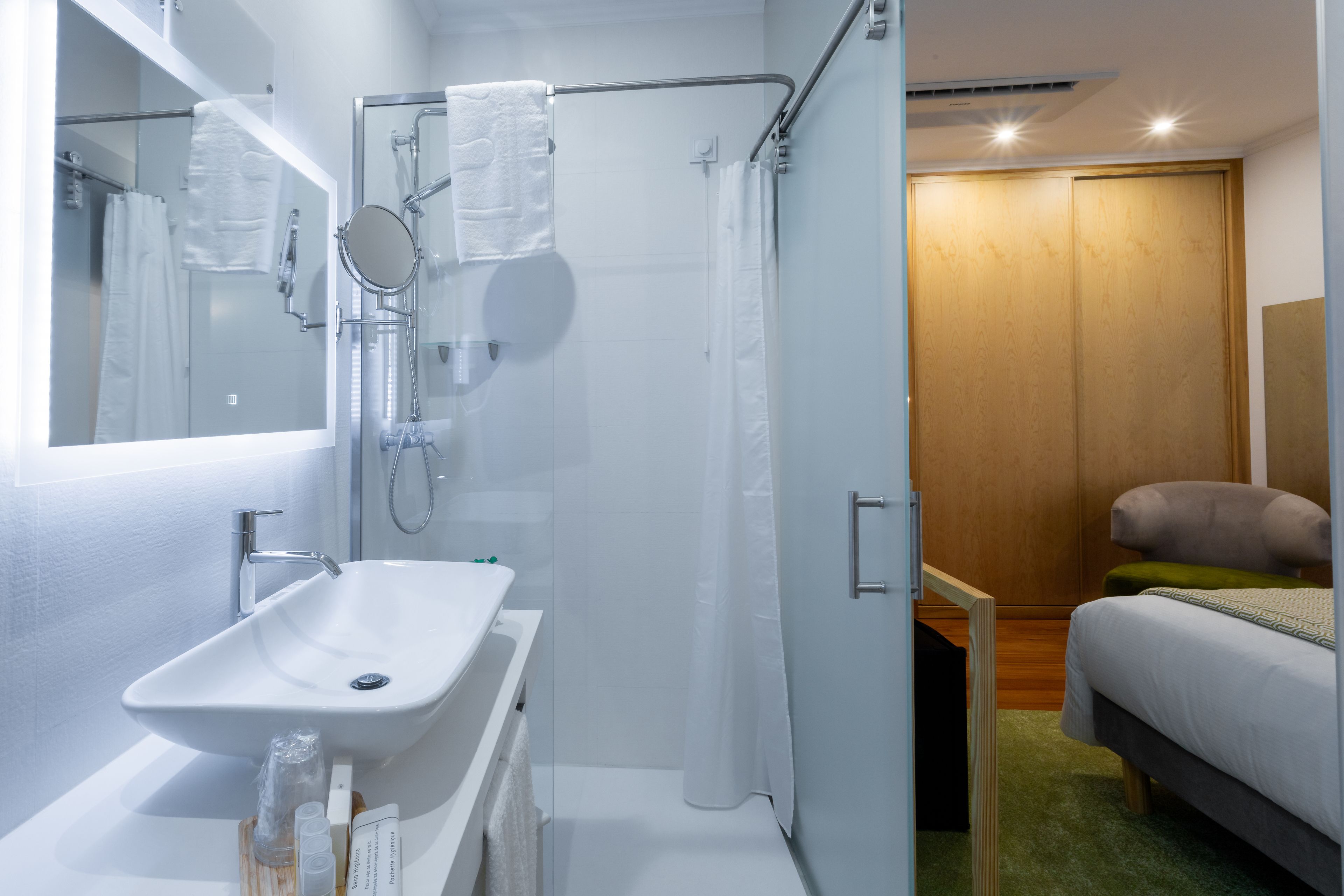 A modern bathroom with a white sink, glass shower enclosure, and a wooden door leading to a bedroom with a bed and a green rug.