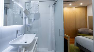 A modern bathroom with a white sink, glass shower enclosure, and a wooden door leading to a bedroom with a bed and a green rug.