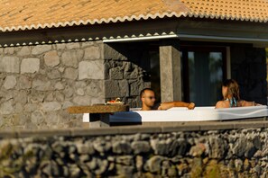 A stone building with a tiled roof, a stone wall, and a hot tub with two people.