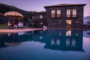 A stone building with a pool, two lounge chairs, and a thatched umbrella.