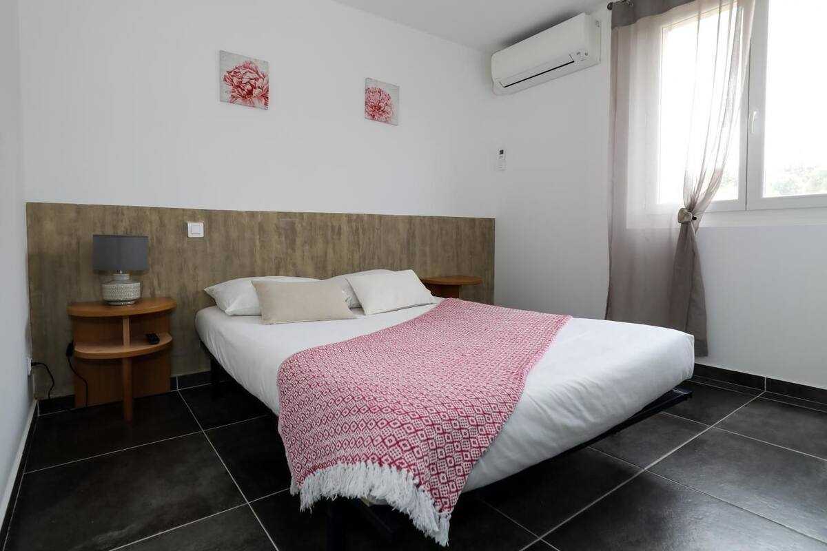 A bedroom with a bed, two bedside tables, a lamp, an air conditioner, and two floral wall art pieces.