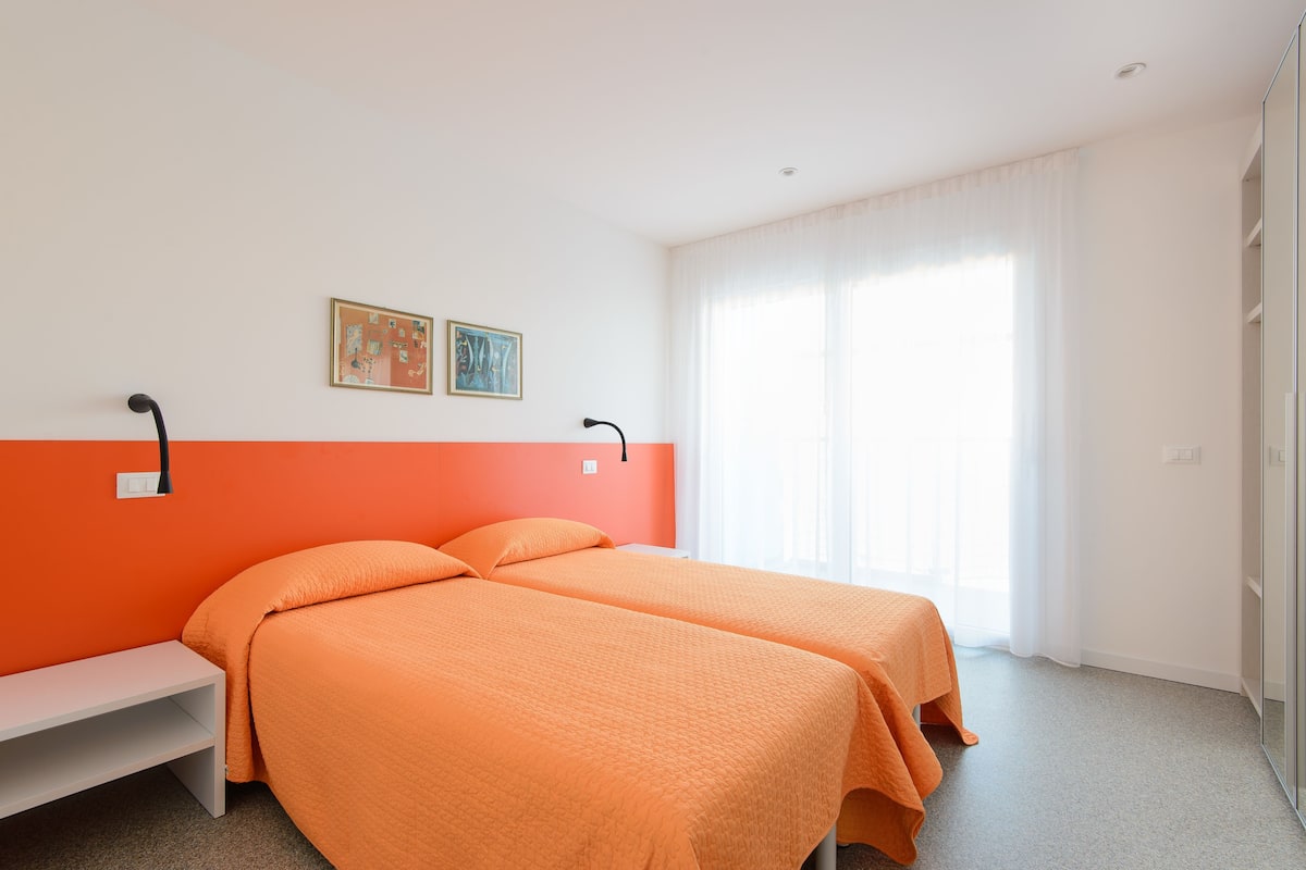 A hotel room with two beds, a bench, a painting, and a large window with curtains.