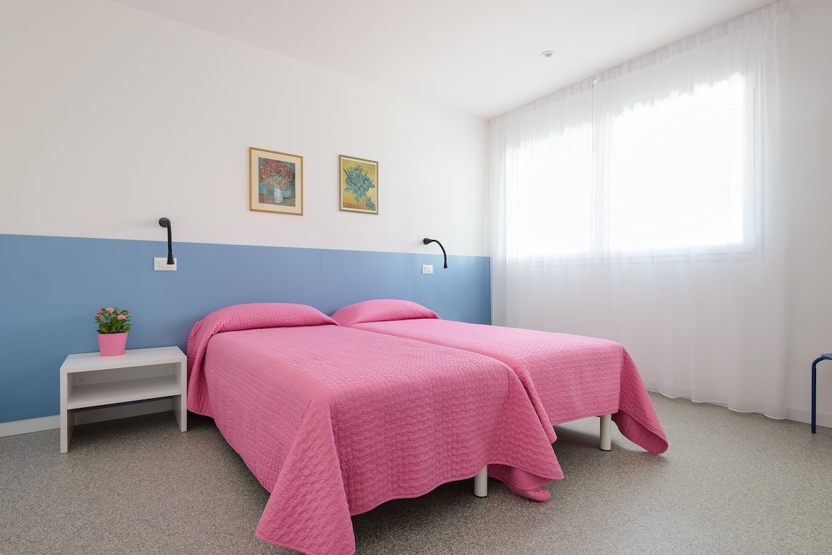 A room with two beds, a nightstand with a pink plant, and two framed pictures on the wall.