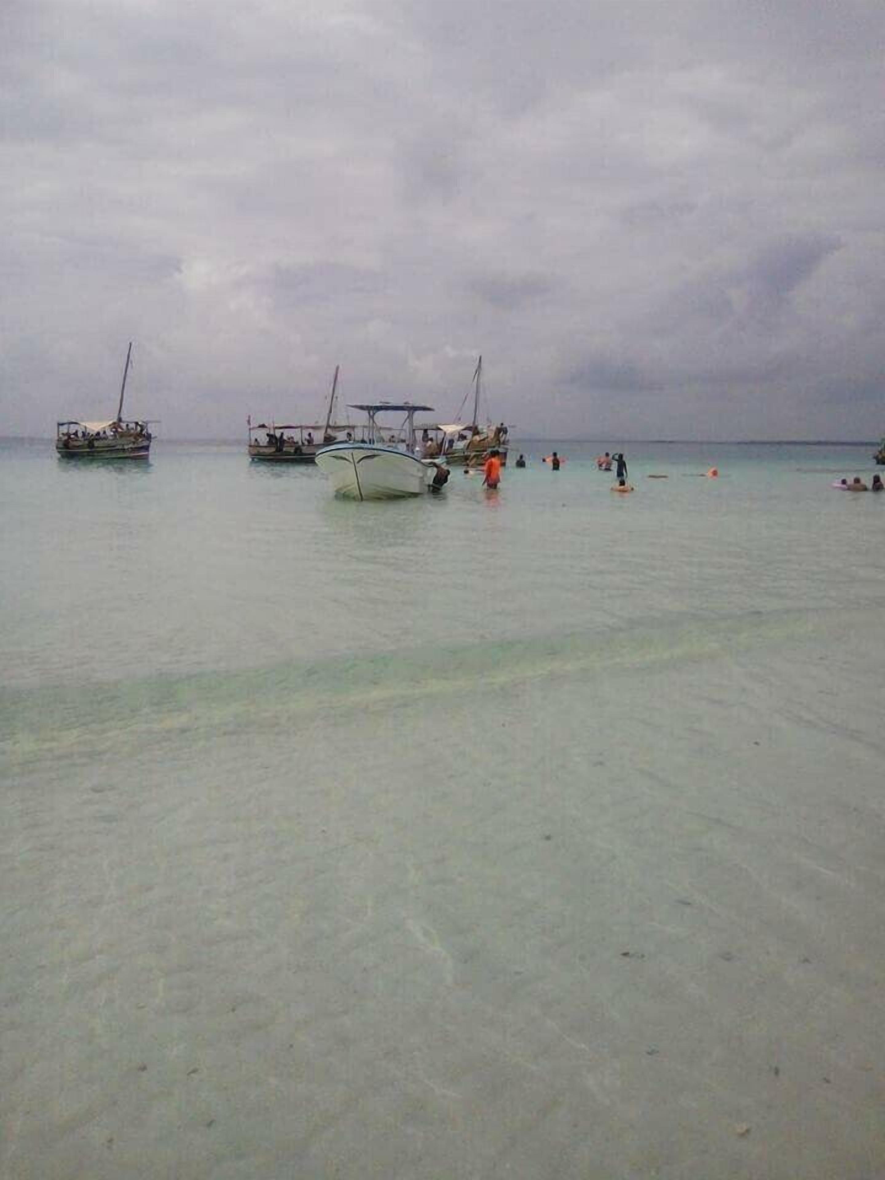 En strand med flere både forankret i rolige farvande, folk svømmer og går langs kysten.