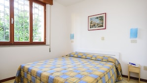 A bedroom with a bed, a window, a painting on the wall, and a small table with books. - Villa Erica (Lignano Sabbiadoro)