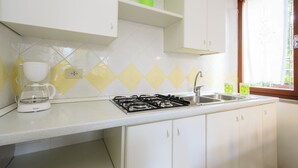A kitchen with white cabinets, a gas stove, a coffee maker, and a window with a view of greenery. - Villa Erica (Lignano Sabbiadoro)