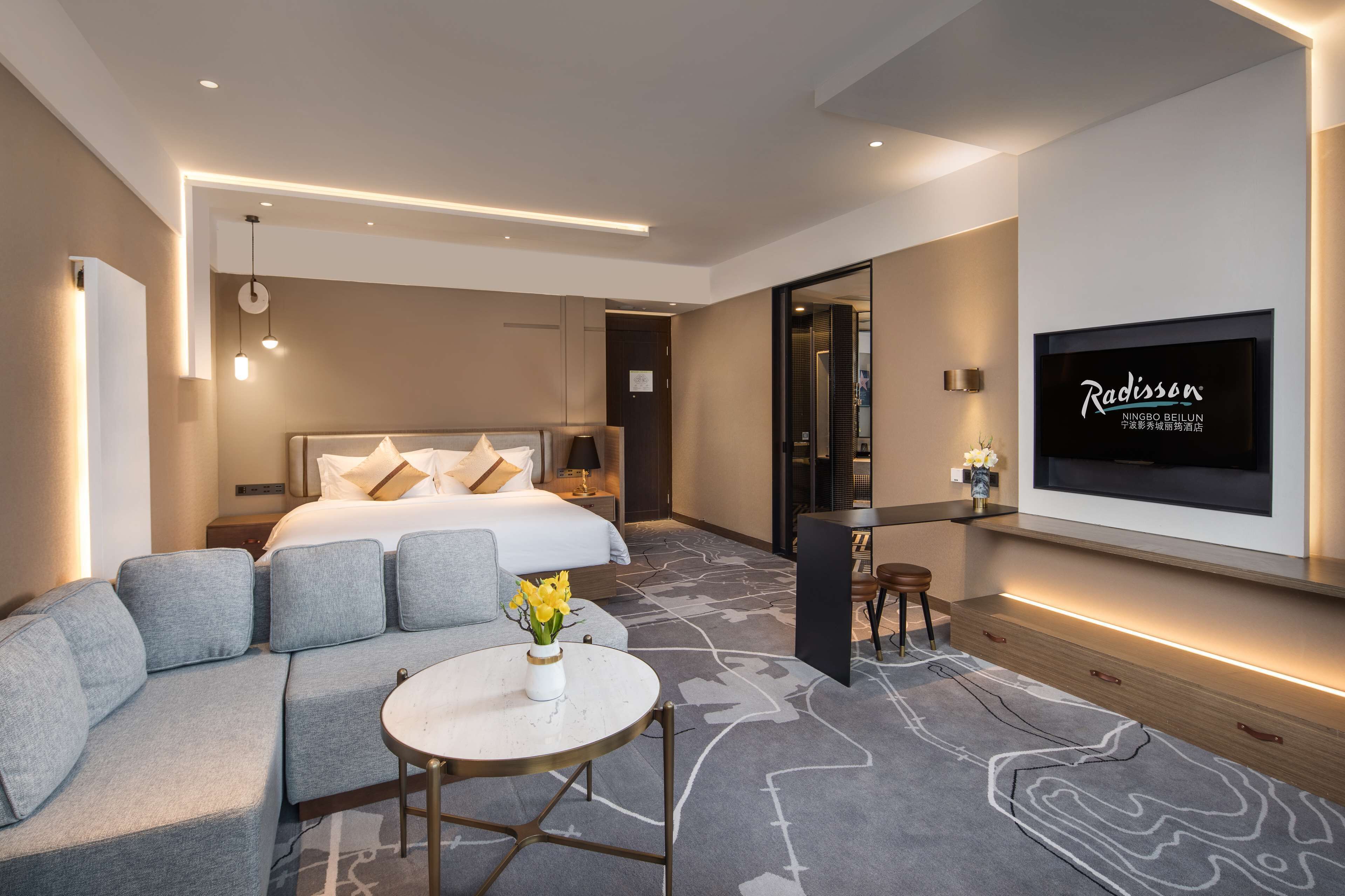 Moderni hotellihuone, jossa on suuri sänky, sohva, pieni pöytä maljakko täynnä kukkia, seinälle asennettu televisio sekä työpöytä ja tuoli.