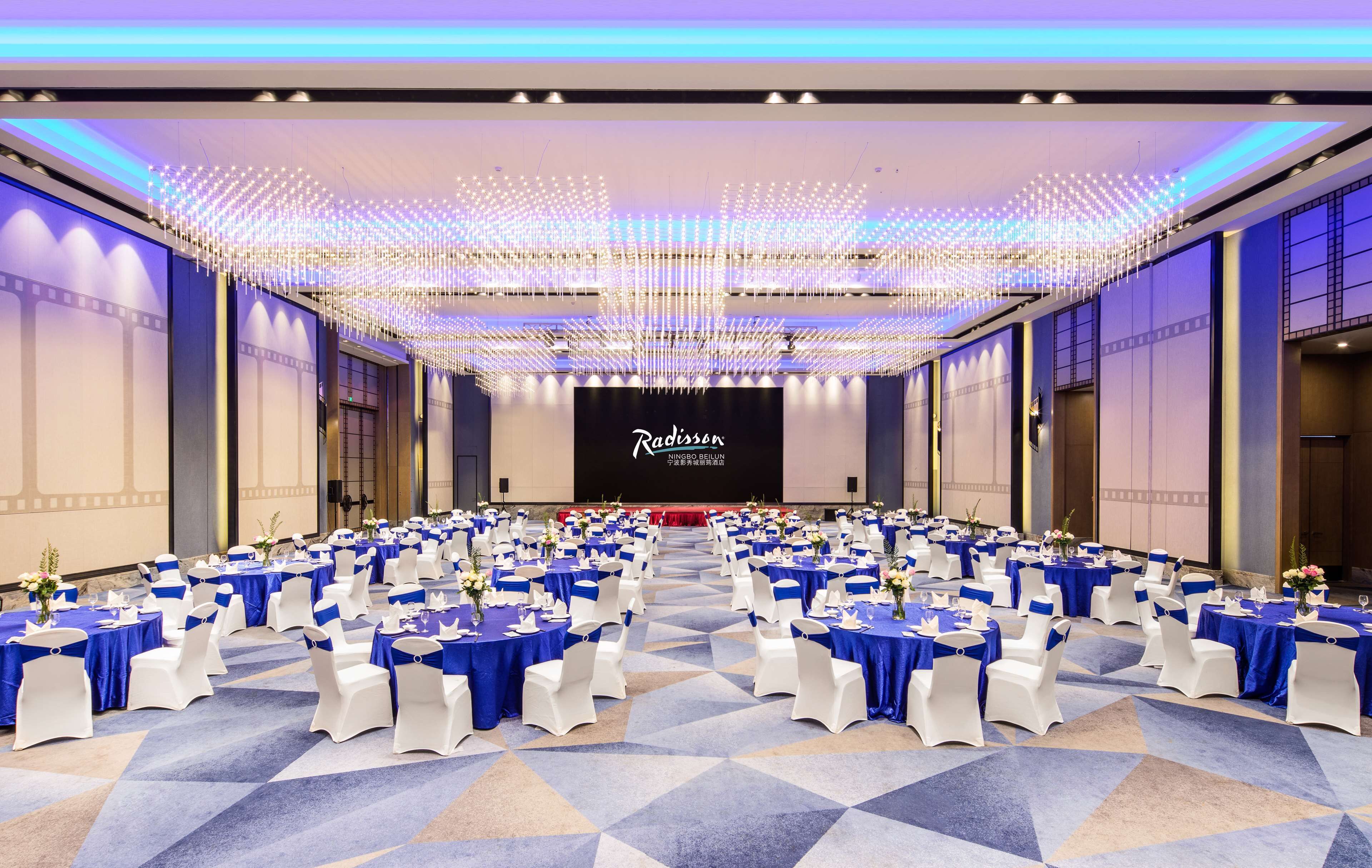 A large conference room with rows of tables and chairs, a stage with a screen, and a ceiling with hanging lights.