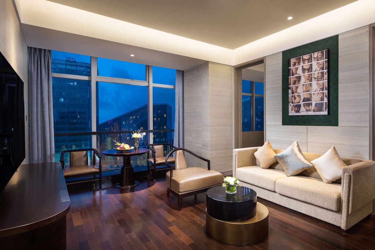 A modern hotel room with a large window offering a city view, a sofa with decorative pillows, a round coffee table, and a television.