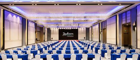 A conference room with rows of tables and chairs, a large screen at the front, and a ceiling with hanging lights.