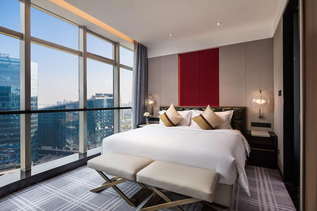 A modern hotel room with a large bed, a bench, and a city view through floor-to-ceiling windows.