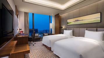 A modern hotel room with two beds, a desk, a TV, and a city view.